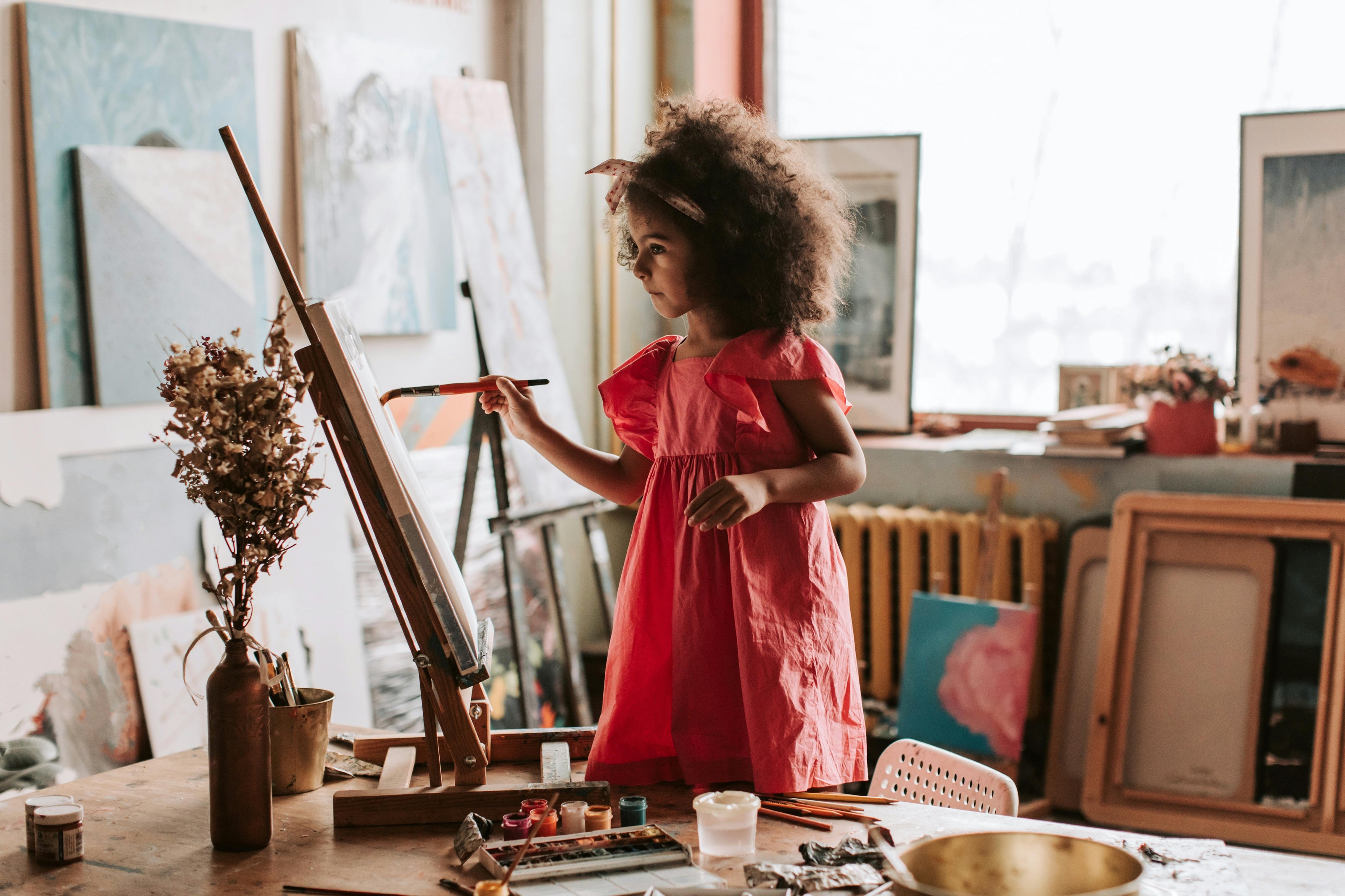 A little girl in a pink dress standing on chair to reach the canvas she is painting