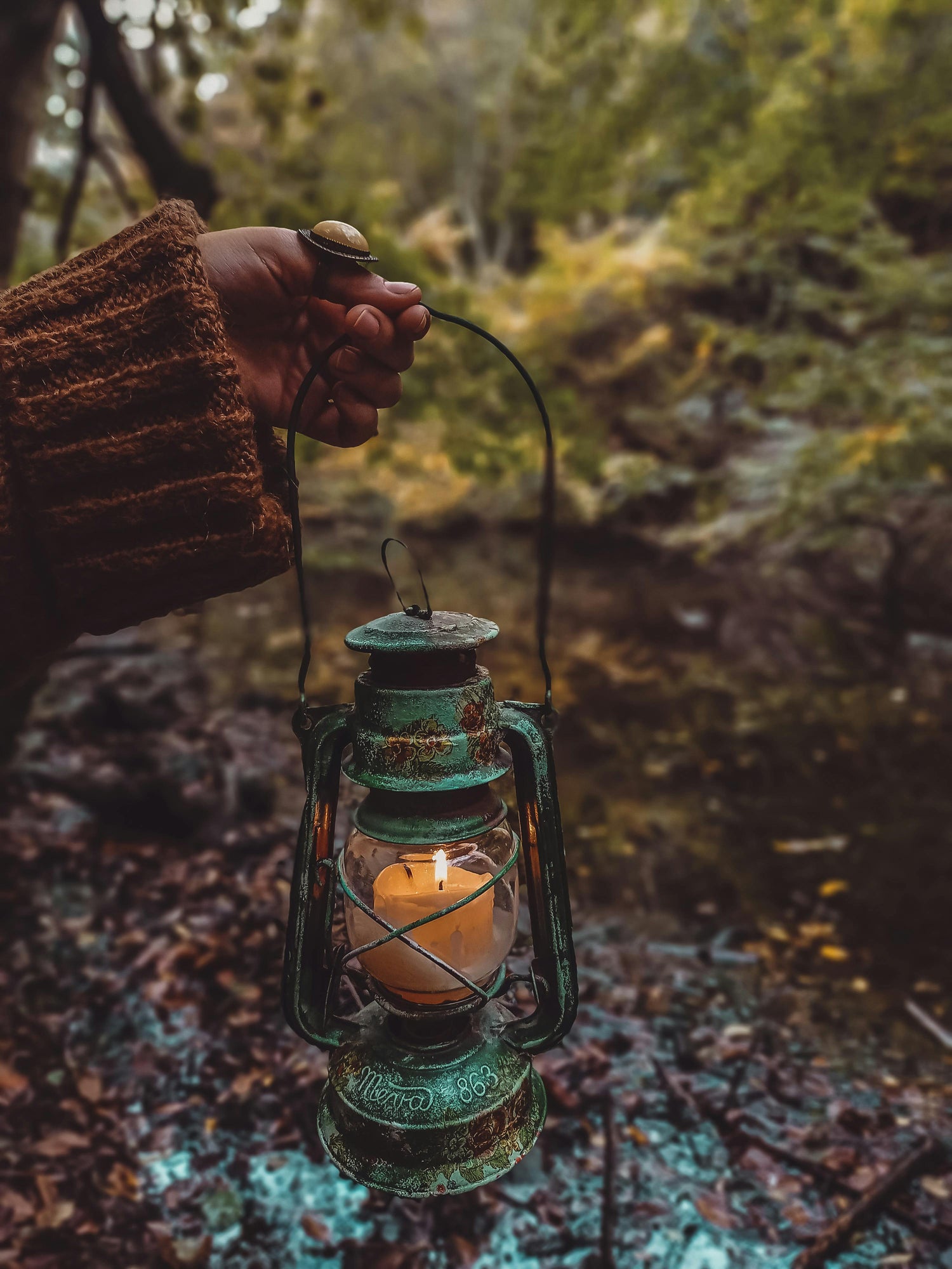 Woman's hand holding a vintage green lantern by a fall river, wearing a burnt orange sweater, symbolizing a personal growth and healing journey.