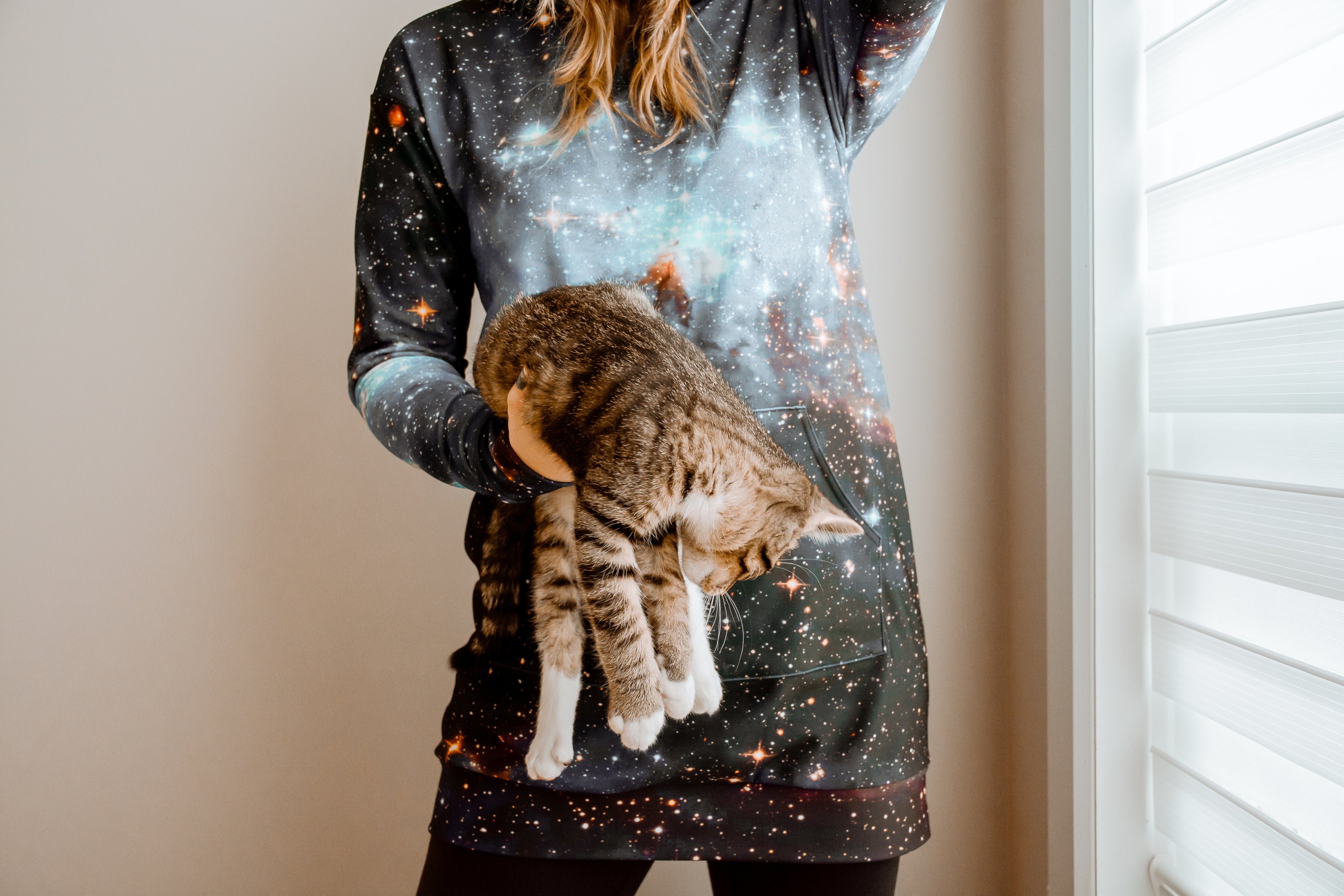 A woman wearing a hoodie holds a cat in one hand next to a window